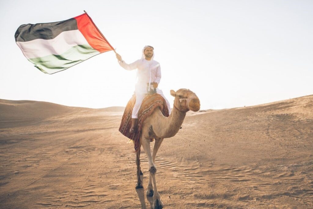 राष्ट्रीय दिवस समारोहों के दौरान गर्व से यूएई का झंडा पकड़े हुए ऊंट की सवारी करता एक अमीराती व्यक्ति पारंपरिक कंदूरा में, दुबई में प्रामाणिक सांस्कृतिक विरासत समारोहों का प्रतिनिधित्व करता है