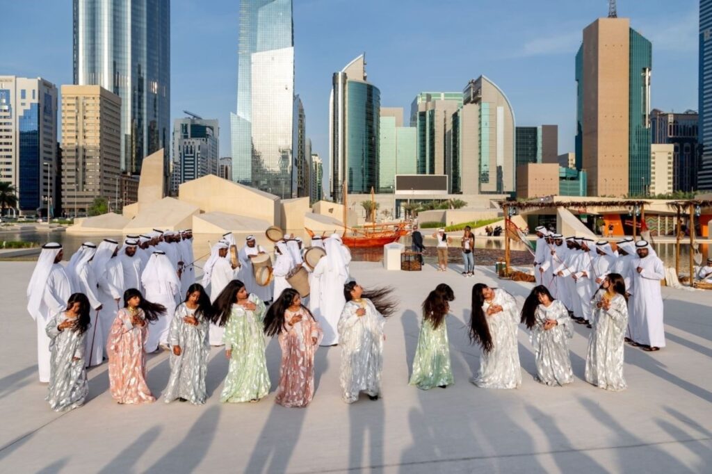 राष्ट्रीय दिवस समारोहों के दौरान रंगीन सांस्कृतिक पोशाक में पारंपरिक अल अयाला नृत्य करते अमीराती पुरुष और युवा लड़कियां, दुबई में प्रामाणिक यूएई विरासत समारोहों का प्रदर्शन करते हुए