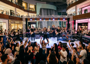 Live music performance in a Dubai Mall atrium with a band on stage, cheering crowd, and luxury storefronts visible.