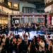 Dubai Mall के एट्रियम में लाइव म्यूजिक परफॉर्मेंस, स्टेज पर बैंड, भीड़ का शोर और विलासवान दुकानें दिख रही हैं।