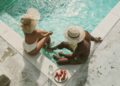 Two people in swimsuits sit at the edge of a turquoise pool, wearing wide-brimmed hats, with a plate of strawberries and a water bottle nearby on the pool deck.