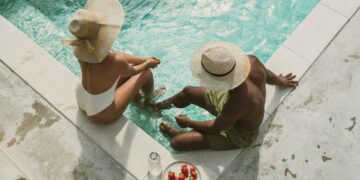 Two people in swimsuits sit at the edge of a turquoise pool, wearing wide-brimmed hats, with a plate of strawberries and a water bottle nearby on the pool deck.