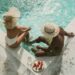 dubai discounts summer dining and spa guide 1 - DUBAIwebzine Two people in swimsuits sit at the edge of a turquoise pool, wearing wide-brimmed hats, with a plate of strawberries and a water bottle nearby on the pool deck.