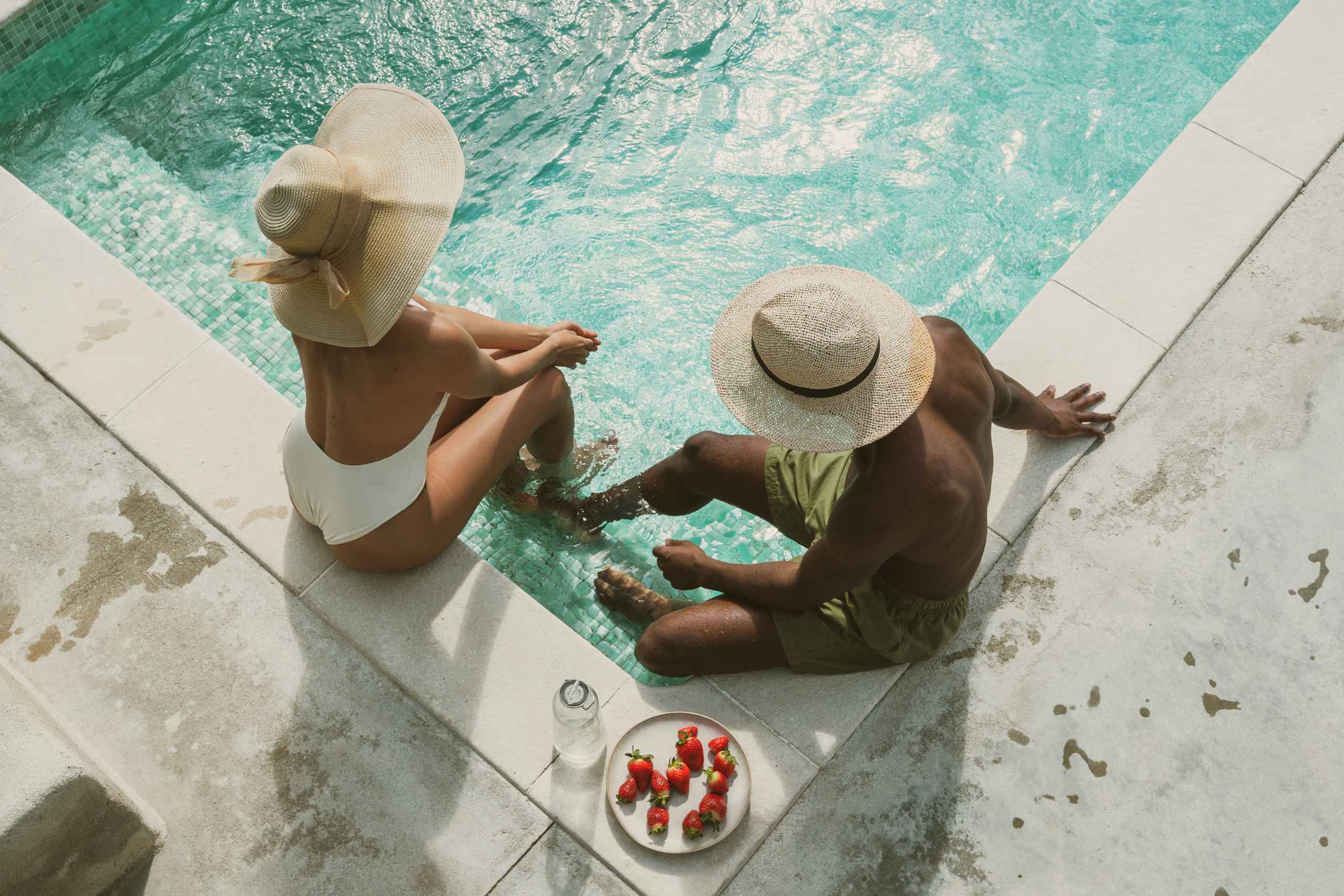 Dos personas en traje de baño sentadas al borde de una piscina turquesa, con sombreros de ala ancha, con un plato de fresas y una botella de agua cerca en la terraza de la piscina.